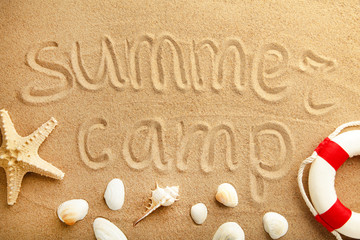 Inscription summer camp on beach sand with seashells and lifebuoy.