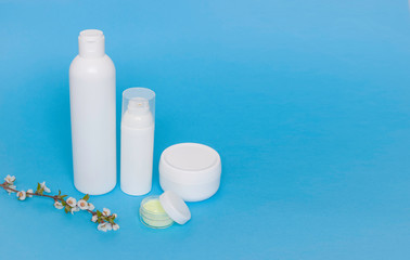 Face creams on a blue background, side view. Twig with flowers in the foreground. Caring for the skin. Free space for text.