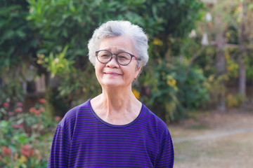 A portrait of an elderly woman wearing eyeglasses, smiling and looking at the camera while standing in a garden. Space for text. Concept of old people and healthcare