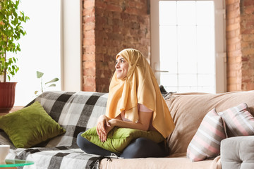 Obraz premium A pretty young muslim woman at home during quarantine and self-insulation. Arabian female model spending time useful being isolated. Concept of healthcare, communication, education during pandemic.