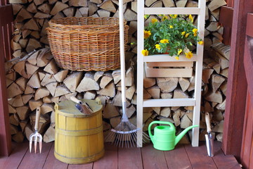 The set of garden tools and bush of Petunia flowers in a wooden shed on the background of stack birch firewood.  - obrazy, fototapety, plakaty