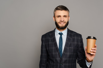 smiling handsome bearded businessman in suit with coffee to go on grey background