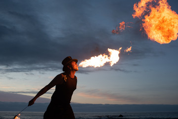 Young man with hat doing Fire breathing on the beach at dusk with sunset in the background.