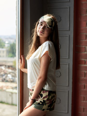 Young brunette woman, wearing jersey pajamas, standing neat big window in dark room in the morning. Hen party celebration. Fashion photography. Bridesmaid on wedding morning.