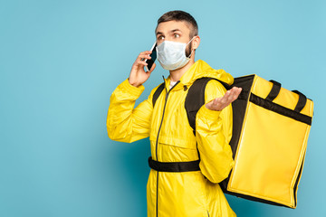 confused deliveryman in medical mask and yellow uniform with backpack talking on smartphone on blue background