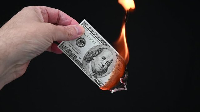 Burning a US $100 bill. A hand holds the banknote while it is on fire. Once it is nearly burned, it is dropped out of the scene. Black background, slow motion.