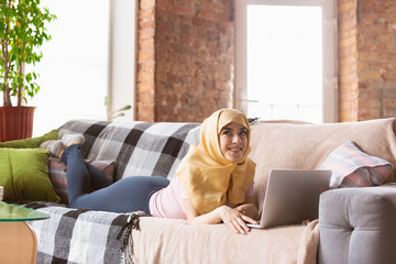 A pretty young muslim woman at home during quarantine and self-insulation. Arabian female model spending time useful being isolated. Concept of healthcare, communication, education during pandemic.