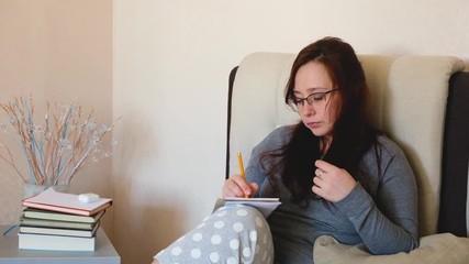 Young caucasian woman writes creative ideas with a pencil.  Brunette girl in glasses writing down her plans in the planner or notes in the notebook. At home.