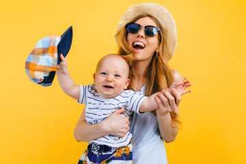 Happy beautiful mother in a hat and sunglasses, holds a little child, on an isolated yellow...