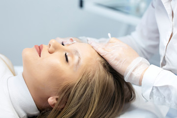 Obraz na płótnie Canvas A young woman lies and master in a white uniform does eyebrow makeup in a beauty salon. The use of permanent makeup on brows. The wizard works with eyebrows. Semi-permanent makeup. Salon background.