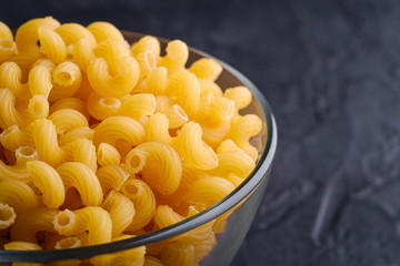 Glass bowl with cavatappi uncooked golden wheat curly pasta on textured dark black background, angle view macro