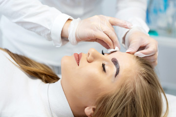 Naklejka premium A young woman lies and master in a white uniform does eyebrow makeup in a beauty salon. The use of permanent makeup on brows. The wizard works with eyebrows. Semi-permanent makeup. Close up