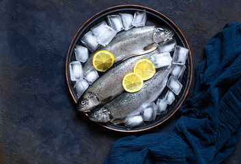 Three fresh raw trouts in ice on a vintage plate with lemons on a dark blue background. Tasty fish ingredient for a healthy dinner