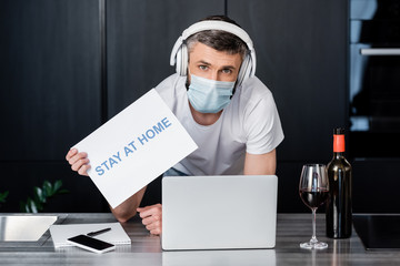 Fototapeta na wymiar Man in medical mask and headphones holding card with stay at home lettering near wine and gadgets on worktop in kitchen