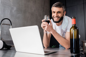 Selective focus of smiling man in medical mask holding glass of wine near laptop and vr headset on worktop in kitchen