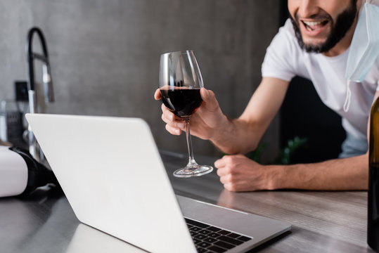 Cropped view of smiling man in medical mask holding glass of wine near laptop on kitchen worktop