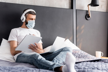 Selective focus of teleworker in headphones and medical mask whiting on notebook and using laptop in bedroom