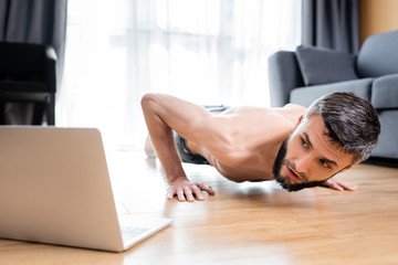 Obraz na płótnie Canvas Selective focus of shirtless sportsman doing push ups near laptop on floor