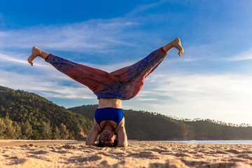 Asian Thai woman practicing yoga at evening in Mae Haad beach, Koh Phangan island, Thailand. Headstand supported pose with legs open.