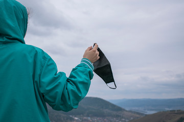 Girl in a birch jacket with a hood holds in her hand a black hygiene mask. View from back....