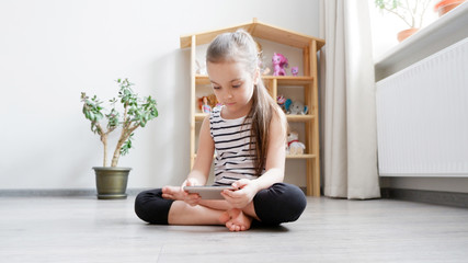 Distance learning. Little girl sitting on a wooden floor with a phone, watching a cartoon or making a video call on a computer
