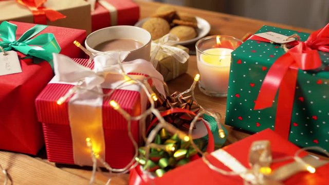 holidays, new year and christmas concept - gift boxes with name tags, candle, hot chocolate drink in mug and electric garland lights on wooden table