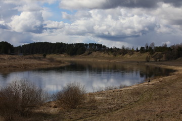 Water landscape in the countryside with wooden houses on a high Bank.A bend in a wide, calm river with a fisherman sitting in the distance with a fishing rod under a cloudy blue sky in spring.Russia