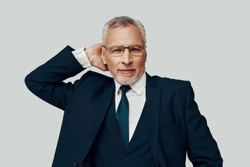 Handsome senior man in full suit looking at camera and smiling while standing against grey background