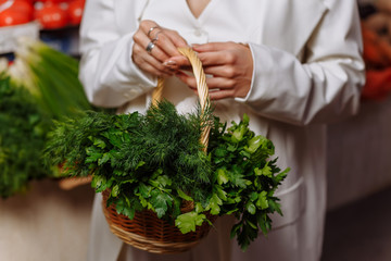 Close-up, a girl holds a wicker basket with fresh herbs in the market at the background of a counter with vegetables.