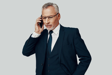 Handsome senior man in full suit talking on the phone while standing against grey background