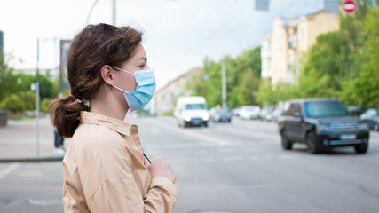 Fototapeta premium Covid-19 pandemic coronavirus concept. Young beautiful curly woman in protective medical face mask standing on street by the road. Girl in face respirator in summer urban city. 