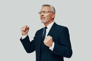 Handsome senior man in full suit gesturing and cheering while standing against grey background