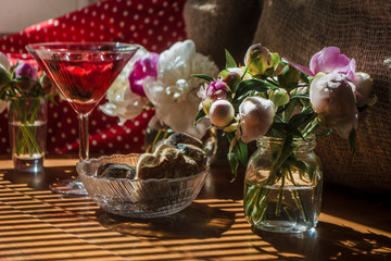 Solemn rustic still life with glass of red drink with cakes in a bowl surrounded by white flowers on textile background on wooden table. Diagonal striped shadow of the blinds on bright sunny day