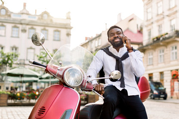 Fototapeta na wymiar Handsome bearded african businessman enjoying his successful phone conversation, while sitting on the modern red motor bike on the background of old city buildings. Business concept