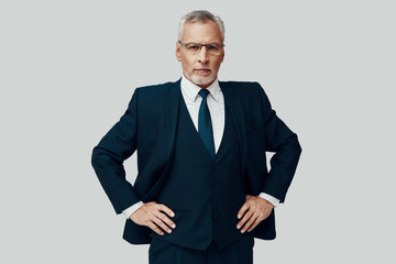 Handsome senior man in full suit looking at camera and keeping hands on hips while standing against grey background