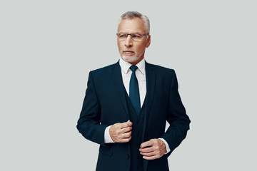 Handsome senior man in full suit looking away and adjusting his suit while standing against grey background