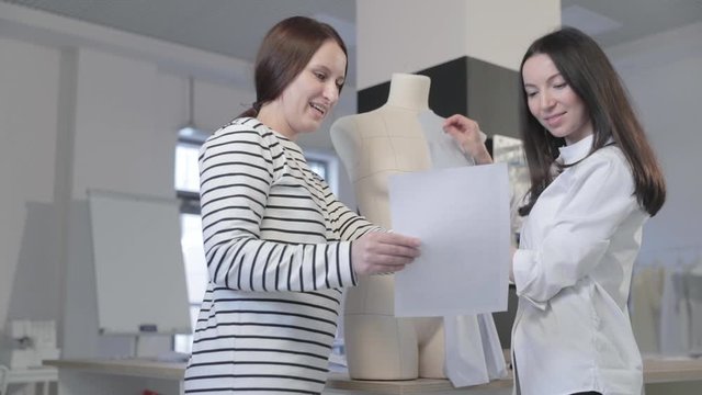Slow motion video of two girls of the fashion designer discuss about something, are looking at a sheet of paper in sewing studio, the sketch of a photo of model in hands, a dummy on background