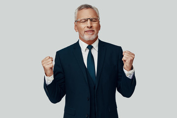 Handsome senior man in full suit gesturing and cheering while standing against grey background