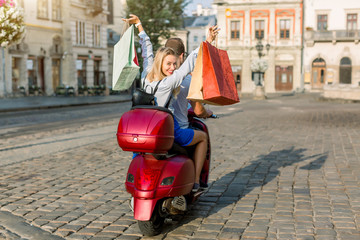 Back view of young couple riding red scooter on a sunny day in old European city. Pretty happy blond woman with shopping bags smiles and looks at camera. Travelling with motor bike, urban tourism © Sofiia Shunkina