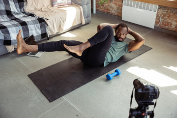 African-american man teaching at home online courses of fitness, aerobic, sporty lifestyle while being quarantine. Activity during insulation, wellness, movement concept. Training, cardio, education.