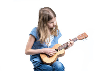 Young, blond girl in blue T-shirt is playing music on ukulele instrument.