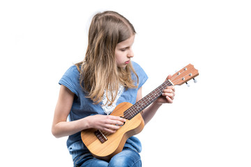 Young, blond girl in blue T-shirt is playing music on ukulele instrument.