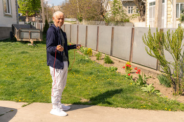 Warming up before workout. Old man in stylish sportswear is jumping with a skipping rope on the outdoor sports ground at home.