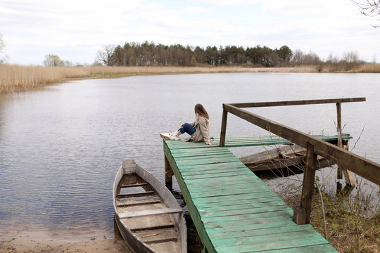 Young beautiful woman sits on the wooden bridge on the river at spring day. old boat near the shore. selective focus.