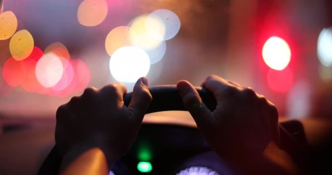 Commuter driver POV holding steering wheel waiting at red light in traffic at night. Green light ON