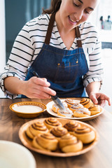 Woman painting freshly baked homemade cinnamon rolls with a delicious cream on a wooden counter in...