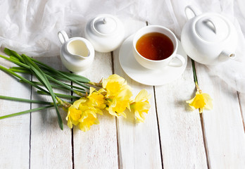 Bunch of yellow daffodils on white wooden background with cup of tea, textile, teapot, milk jug, sugar bowl
