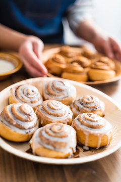 Close up of a woman picking up a plate of freshly baked homemade cinnamon rolls on a wooden counter in her kitchen.