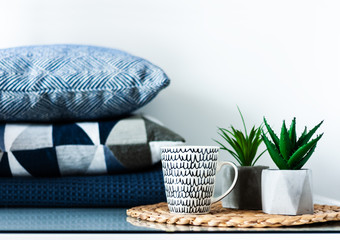 Cozy home interior decor: white and black cup and ornamental plants in pots on a wicker stand on a white table in the room. Blue pillows on background. The quarantine concept of stay home.
