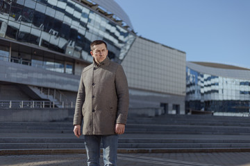 A young adult man in a gray coat walks along a city street on a sunny day in spring